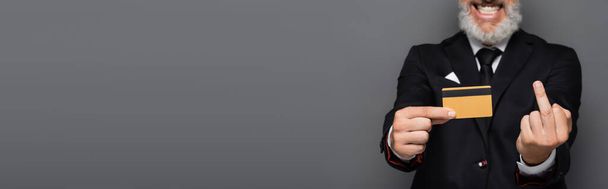 обрізаний вид веселого бізнесмена, що тримає кредитну картку, показуючи середній палець ізольовано на сірому, банер
 - Фото, зображення