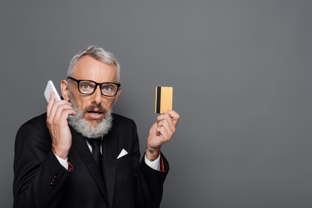 überraschter Geschäftsmann mittleren Alters mit Kreditkarte, während er auf dem Smartphone isoliert auf grau spricht - Foto, Bild
