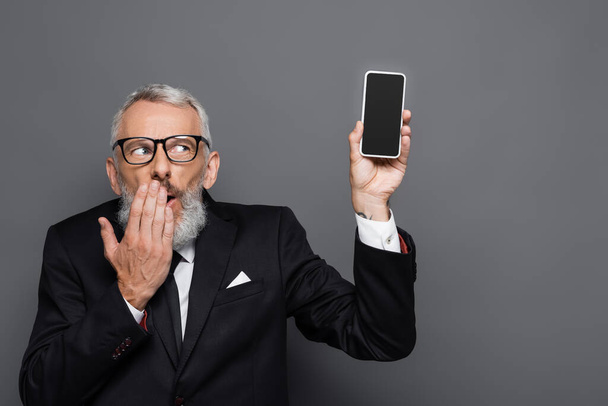 middle aged businessman holding cellphone with blank screen and covering mouth on grey - Fotografie, Obrázek