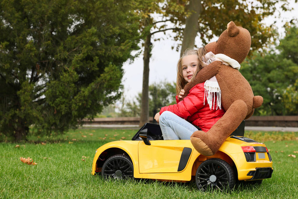 Cute little girl with toy bear driving children's car in park. Space for text - Фото, зображення