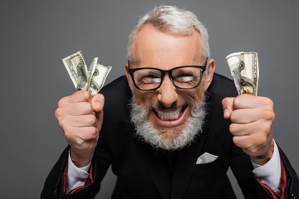 happy middle aged businessman in glasses holding dollar banknotes and grinning isolated on grey - Photo, Image