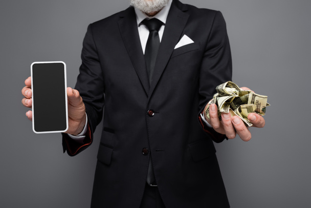 καλλιεργημένη άποψη μεσήλικας επιχειρηματίας που κατέχει τραπεζογραμμάτια και smartphone σε δολάρια με λευκή οθόνη απομονωμένη σε γκρι - Φωτογραφία, εικόνα