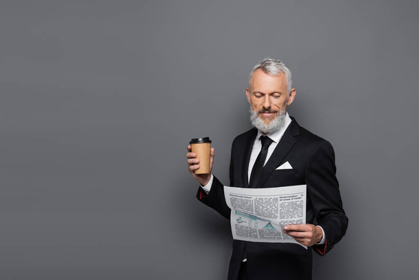 Lächelnder Geschäftsmann mittleren Alters mit Pappbecher und Zeitung auf grau - Foto, Bild
