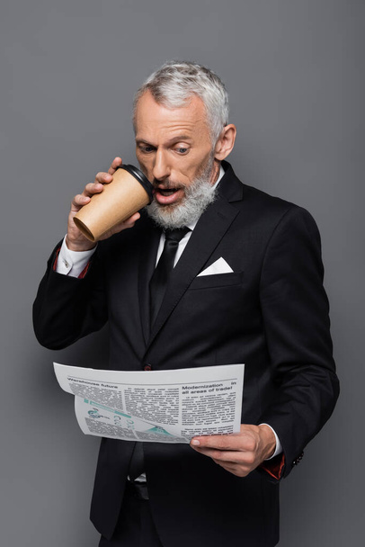 shocked middle aged businessman holding paper cup and reading newspaper on grey - Foto, Bild