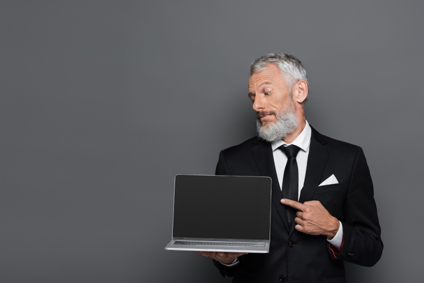 Geschäftsmann mittleren Alters im Anzug zeigt auf Laptop mit leerem Bildschirm auf grau  - Foto, Bild