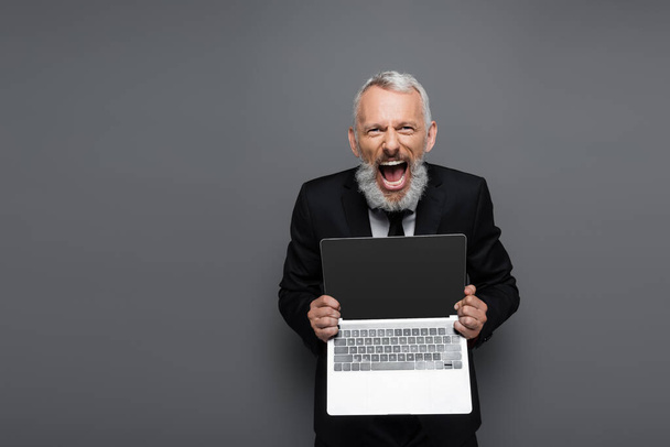 amazed middle aged businessman in suit holding laptop with blank screen on grey  - Fotografie, Obrázek