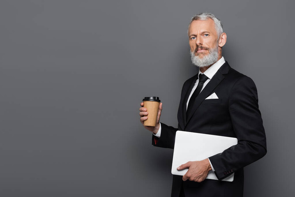 middle aged businessman in suit holding laptop and paper cup on grey  - 写真・画像