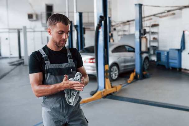 Wiping hands from a dirt. Man in black shirt and grey uniform stands in garage after repairing broken car. - Φωτογραφία, εικόνα