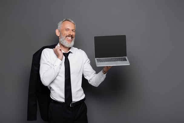 happy middle aged businessman in suit holding laptop with blank screen and blazer on grey  - Photo, Image