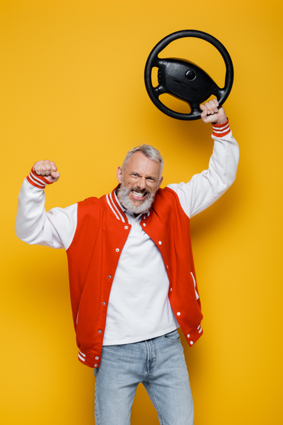 happy middle aged man in bomber jacket holding steering wheel above head on yellow - Zdjęcie, obraz