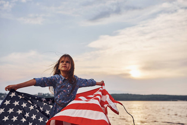 Amazing sunset. Patriotic female kid with American Flag in hands. Against cloudy sky. - Foto, Imagem