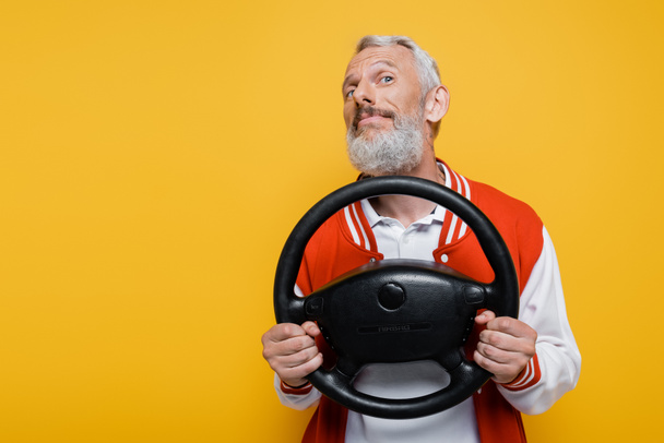 pleased middle aged man in bomber jacket holding steering wheel while imitating driving isolated on yellow - Zdjęcie, obraz