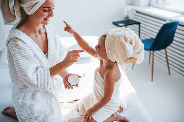 Using cream to clear skin. Young mother with her daugher have beauty day indoors in white room. - Photo, Image