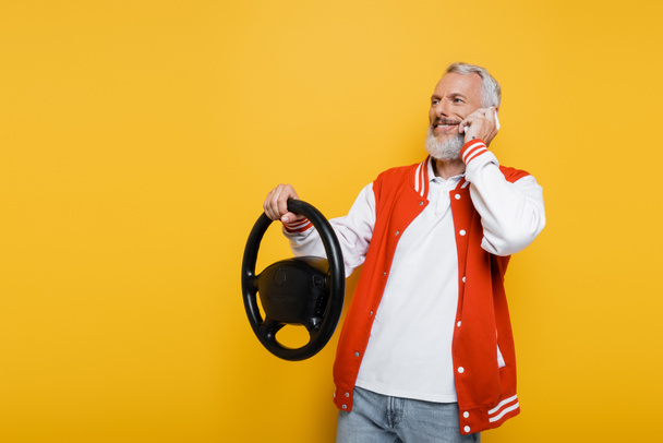 happy middle aged man holding steering wheel while imitating driving and talking on smartphone isolated on yellow - Foto, immagini