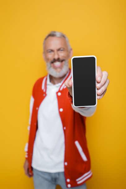 happy and blurred middle aged man holding smartphone with blank screen isolated on yellow  - 写真・画像
