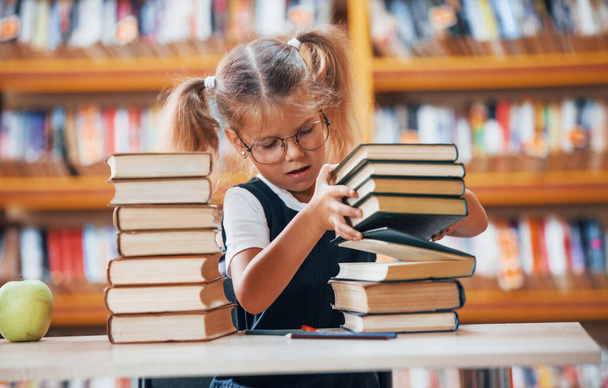 Cute little girl with pigtails is in the library. Apple on the books. - 写真・画像