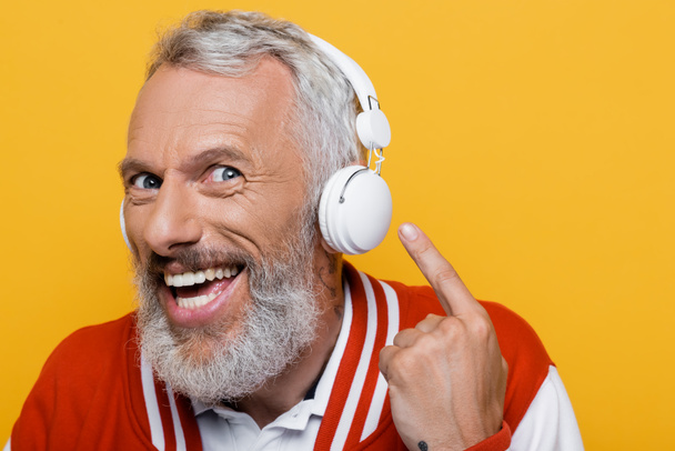 positive middle aged man pointing at wireless headphones and listening music isolated on yellow  - Photo, Image