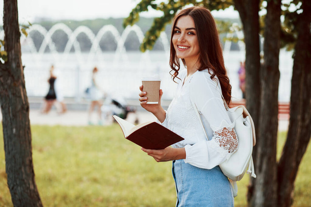Happy girl with book and cup of fresh coffee. Young woman have good time in the park at her weekend. - Foto, afbeelding