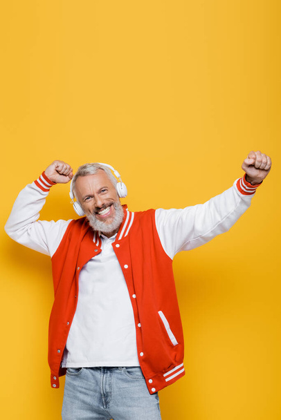 joyful middle aged man in wireless headphones listening music and dancing on yellow  - Photo, Image
