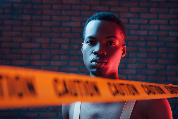 Yellow caution tape and uniform. Futuristic neon lighting. Young african american man in the studio. - Фото, зображення