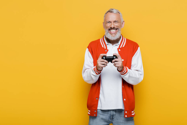 QUIIV, UCRÂNIA - JULHO 29, 2021: homem feliz de meia-idade com casaco de bombista segurando joystick isolado no amarelo - Foto, Imagem