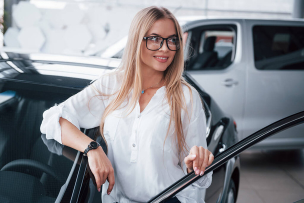 With van behind. Cute girl in eyewear stands near the car in auto saloon. Probably her next purchase. - Fotografie, Obrázek