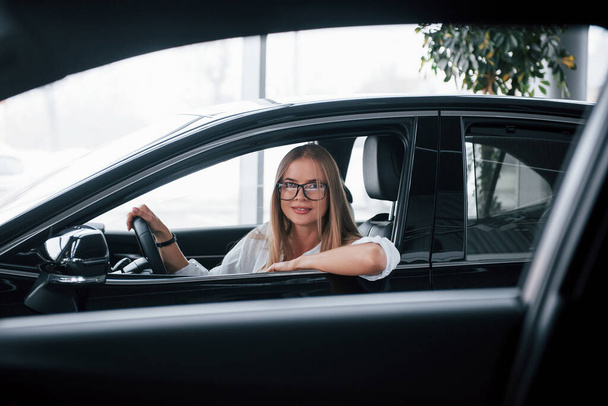 Hand on the opened window. Beautiful blonde girl sitting in the new car with modern black interior. - Valokuva, kuva