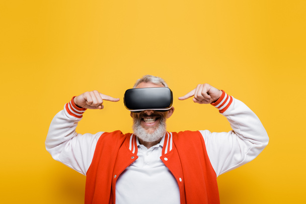 happy middle aged man in bomber jacket pointing at vr headset on yellow - Foto, afbeelding