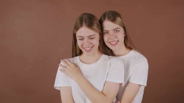 Retrato de hermanas gemelas jóvenes felices en camisetas blancas posando para la cámara sobre fondo marrón - Metraje, vídeo