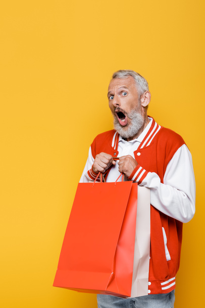shocked middle aged man in bomber jacket holding shopping bags isolated on yellow - Фото, изображение