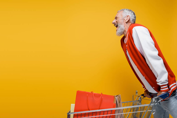 side view of excited middle aged man in bomber jacket near cart with shopping bags isolated on yellow - Fotografie, Obrázek