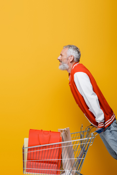 Seitenansicht eines glücklichen Mannes mittleren Alters in Bomberjacke neben Einkaufswagen mit Einkaufstaschen auf gelb - Foto, Bild
