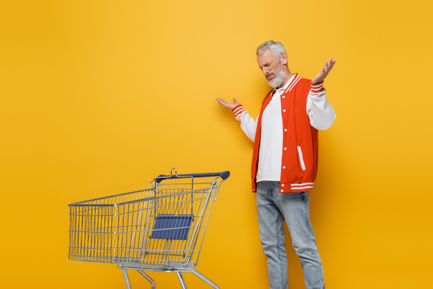 elégedetlen középkorú férfi bombázó kabát mutatja vonós gesztus közelében üres bevásárlókocsi sárga - Fotó, kép