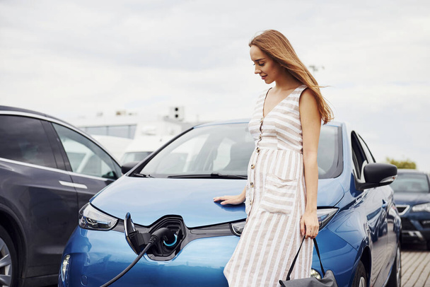 Cloudy day. Woman on the electric cars charge station at daytime. Brand new vehicle. - 写真・画像