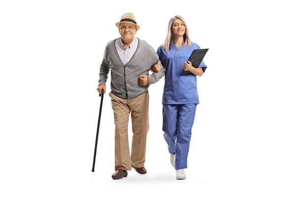 Plně dlouhý portrét mladé ošetřovatelky v modré uniformě, pomáhající staršímu muži izolovanému na bílém pozadí - Fotografie, Obrázek