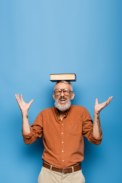tattooed middle aged teacher in glasses and book on head gesturing on blue - 写真・画像