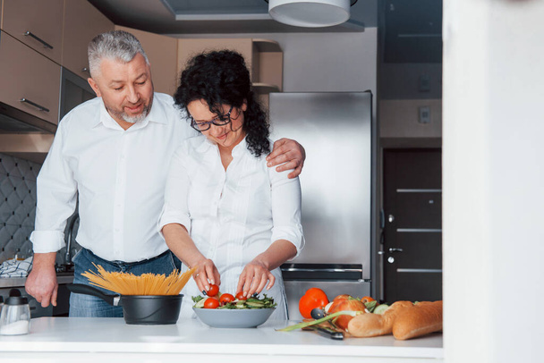 L'amore è come il vino, sempre meglio con ogni anno. Uomo e sua moglie in camicia bianca preparare il cibo in cucina utilizzando verdure. - Foto, immagini