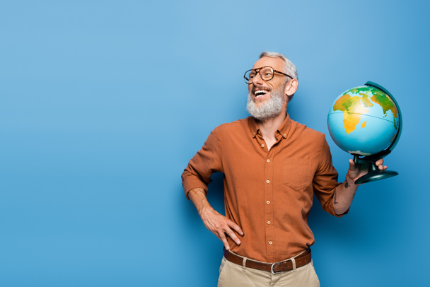 happy middle aged teacher in glasses holding globe and standing with hand on hip on blue - Zdjęcie, obraz