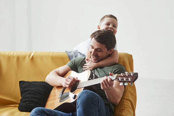 Playful mood. Father and his son sitting on the sofa together. Acoustic guitar in hands. - Foto, afbeelding