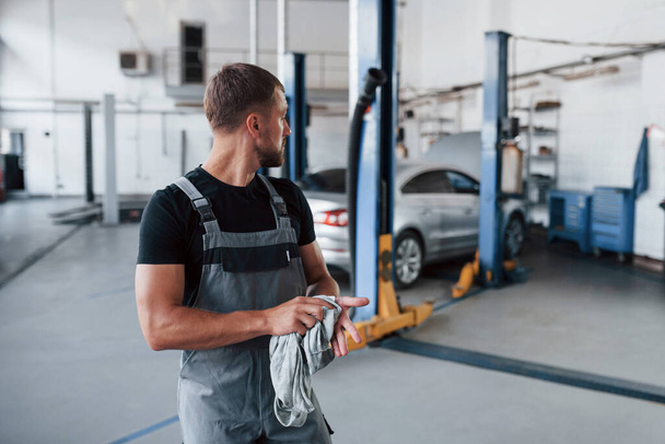 Looks behind. Man in black shirt and grey uniform stands in garage after repairing broken car. - Foto, imagen