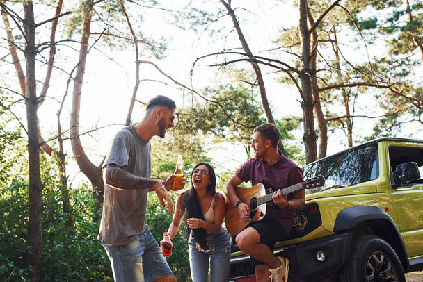 Singing songs together. Friends have nice weekend outdoors near theirs green car with acoustic guitar. - Foto, Imagen