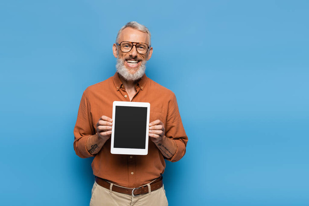 glücklicher Mann mittleren Alters in Brille und Hemd mit digitalem Tablet mit leerem Bildschirm auf blauem Hintergrund - Foto, Bild