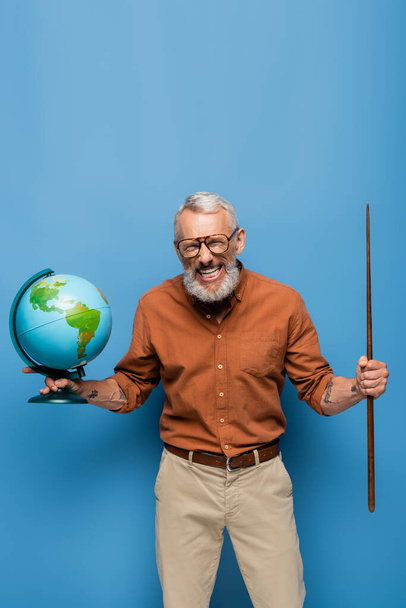 tevreden middelbare leeftijd leraar in bril houden pointer stick en globe op blauw - Foto, afbeelding