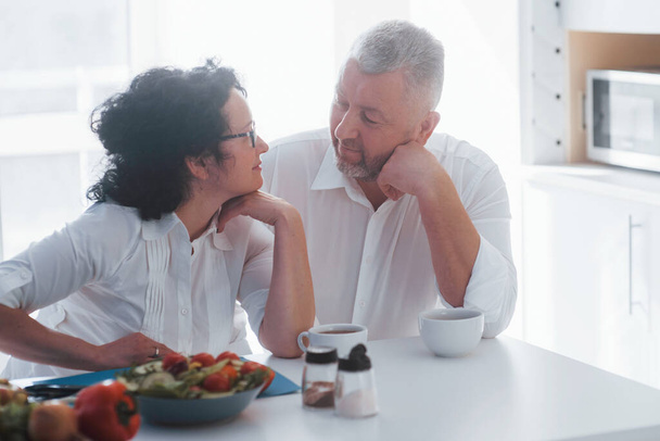 Looking at each other with love. Senior man and his wife in white shirt have romantic dinner on the kitchen. - Fotoğraf, Görsel
