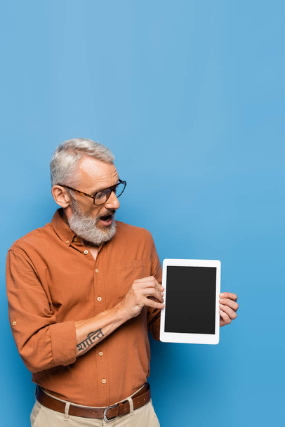 удивленный мужчина средних лет в очках и рубашке, глядя на цифровой планшет с чистым экраном на синем - Фото, изображение