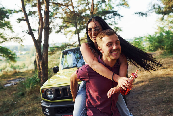 Smiling and having fun. Couple of young people with alcohol have fun in the forest. Green jeep behind. - Фото, изображение