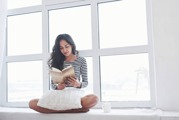 Girl reading book while sitting on the windowsill. Having free day. - Foto, imagen