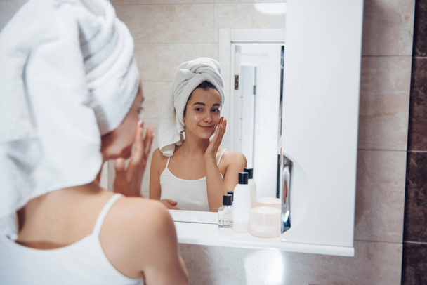 Putting cream on the face. In the bathroom. Woman in white towel in head have care of herself. - Photo, image