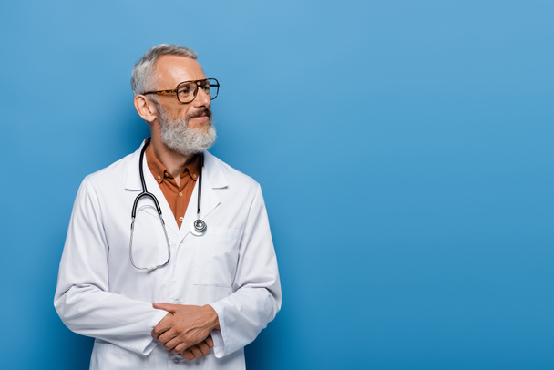 満足している中年の医者で眼鏡と白いコートが青い上に立っている - 写真・画像