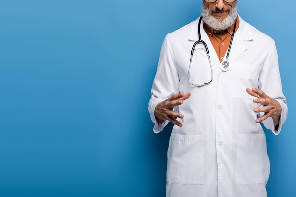 καλλιεργημένη άποψη του γενειοφόρου μεσήλικας γιατρός σε λευκό παλτό gesturing σε μπλε - Φωτογραφία, εικόνα